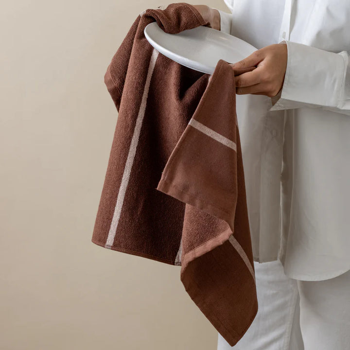 Brown towel with white stripes held by a person wearing a white shirt and pants against a beige background
