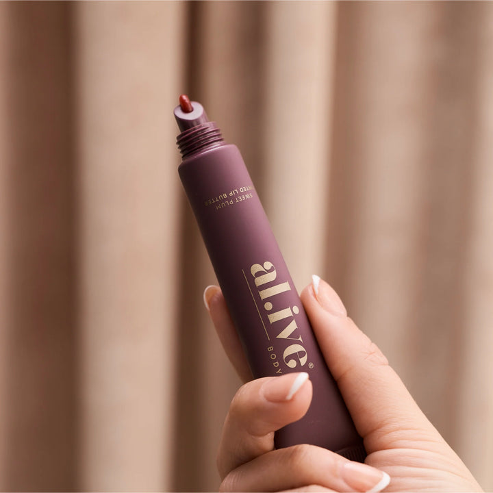 Hand holding a purple cosmetic product with 'alive' branding against a beige background