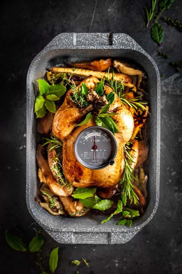 Roasted chicken with herbs in a roasting pan with a meat thermometer on a dark background