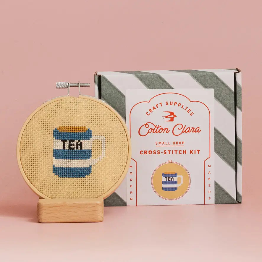 Cross-stitch kit with a completed design of a tea cup on a pink background, featuring the Cotton Clara brand.