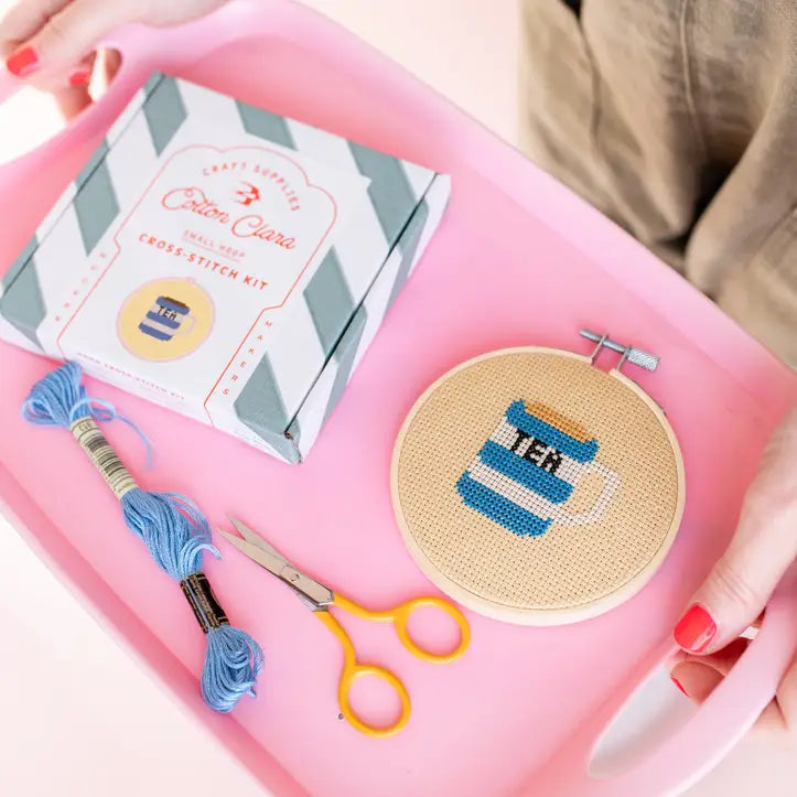 Cross-stitch kit with hoop, threads, scissors, and a completed cross-stitch design on a pink tray.