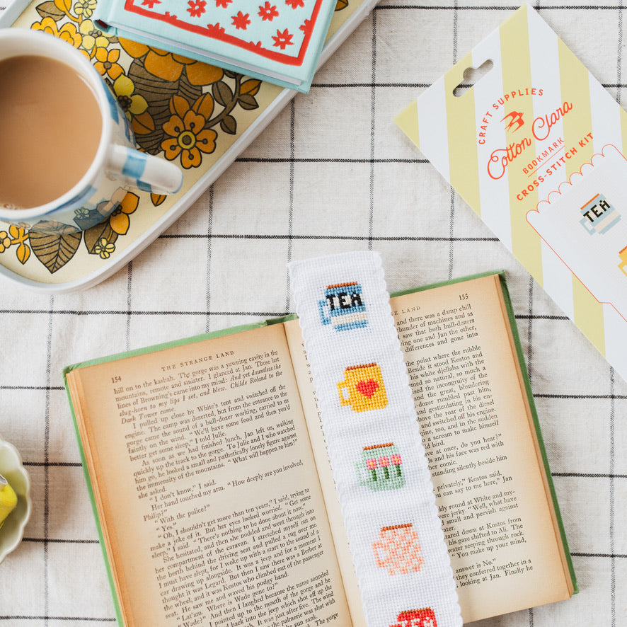 Cross-stitched bookmark with tea-themed designs on an open book, next to a cup of coffee and craft supplies.