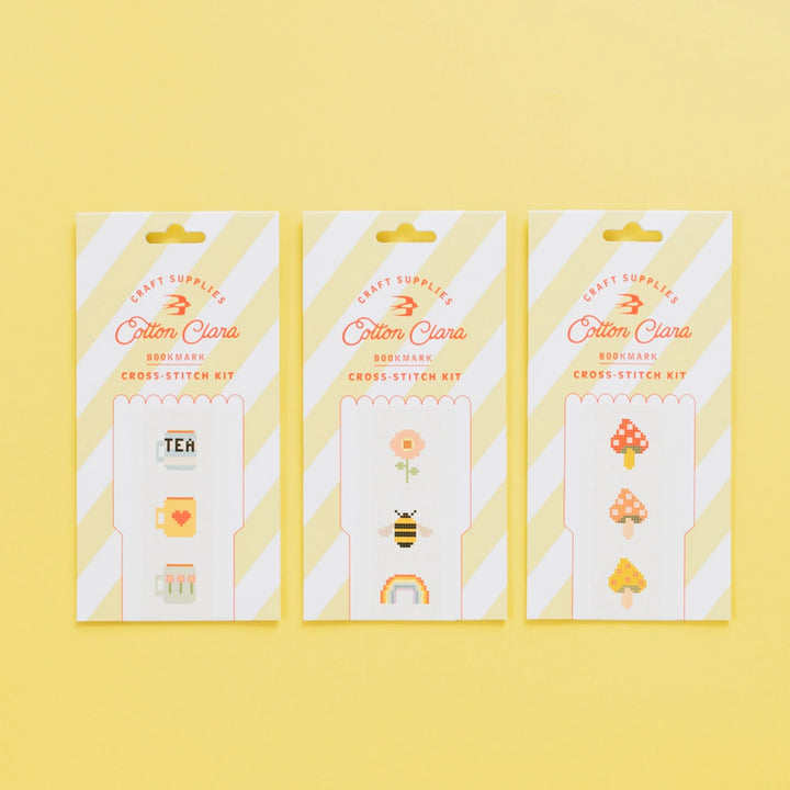 Three packages of Cotton Clara cross-stitch kits on a yellow background