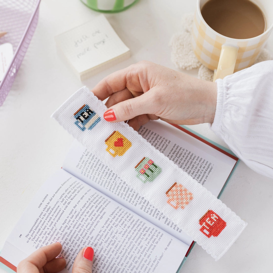 Person holding a cross-stitched bookmark with colorful designs on an open book.