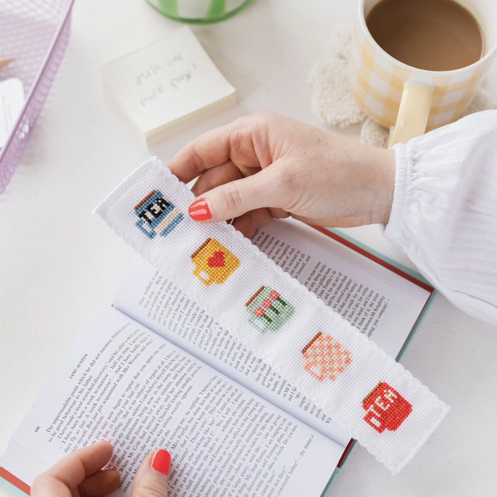 Person holding a cross-stitched bookmark with colorful designs on an open book.
