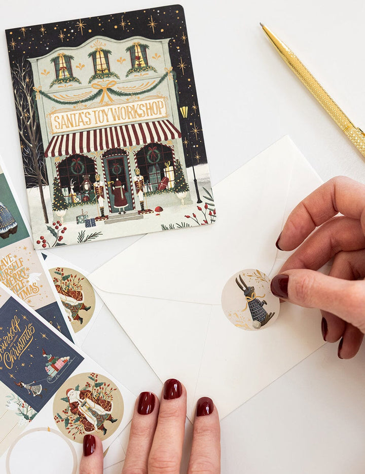 Hand holding a Christmas-themed card with Santa's Workshop design, surrounded by other cards and decorative elements.