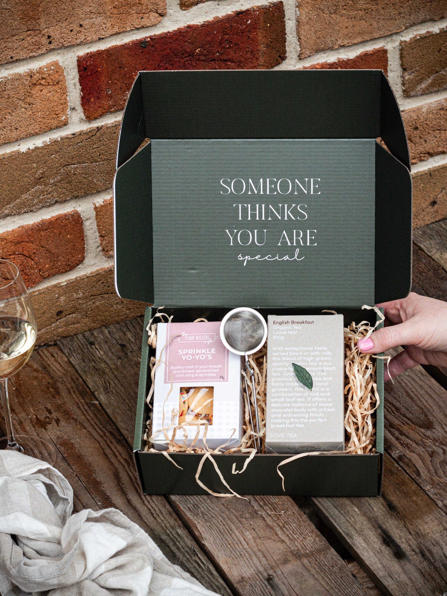 A gift box including biscuits, tea and a tea infuser on a wooden bench with a brick background 