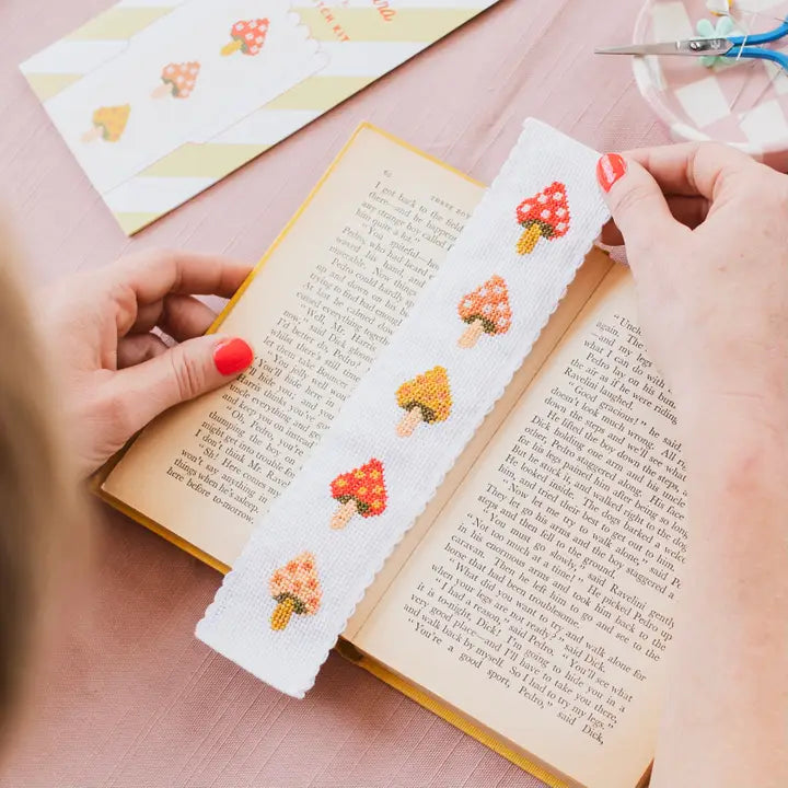 Person holding a cross-stitched bookmark with mushroom designs over an open book.