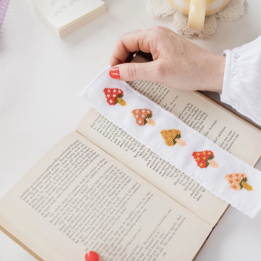 Hand holding a cross-stitched bookmark with mushroom pattern over an open book.