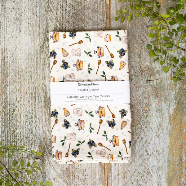 A patterned tea towels on a wooden surface with greenery.