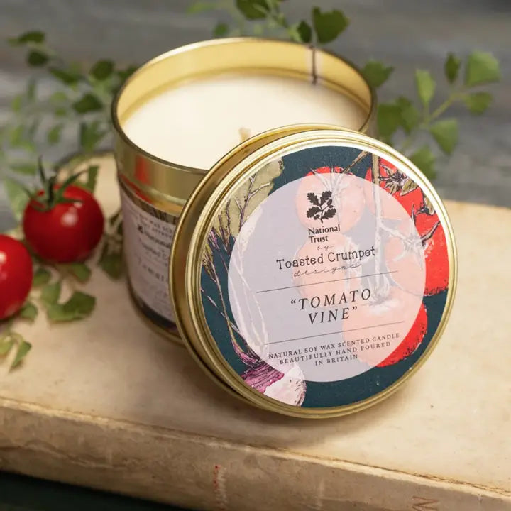 Candle in a tin with 'Tomato Vine' label on a wooden surface with tomatoes and leaves.