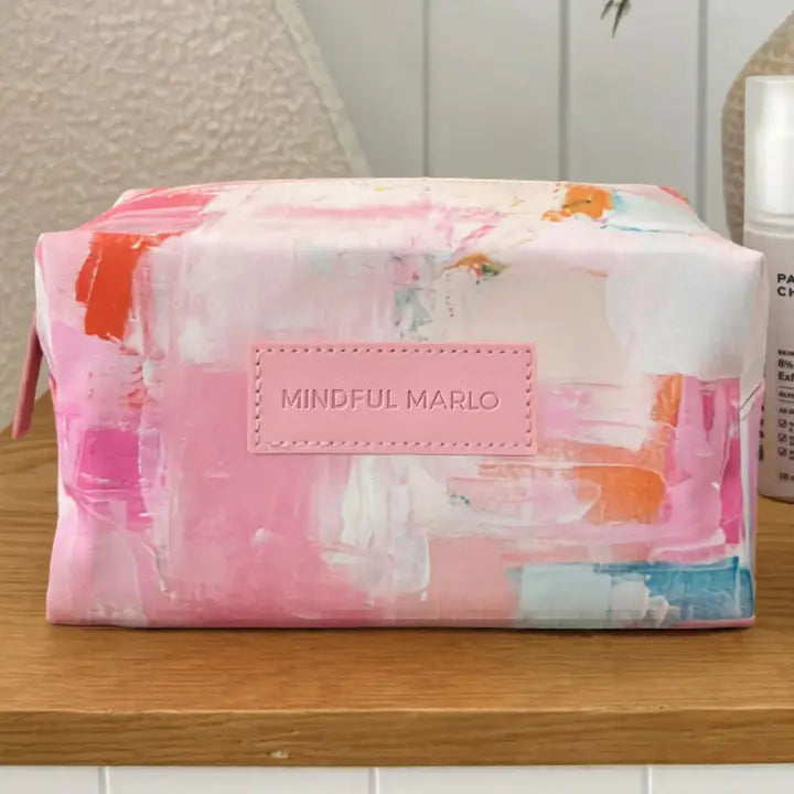 A rectangular makeup bag with a pink and white abstract paint brush stroke pattern sits on a wooden surface. The bag features a central pink patch with the text &