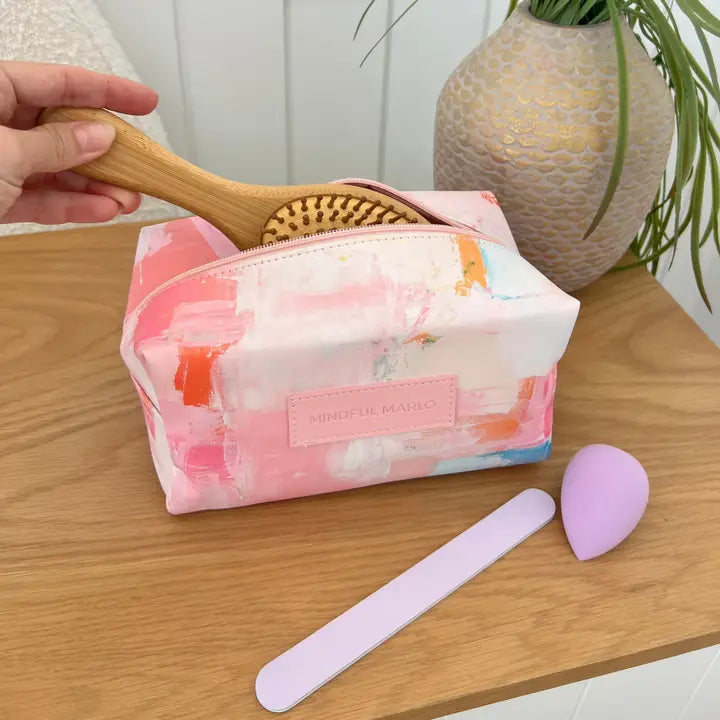 Makeup bag with floral design, wooden brush, and pink spatula on a wooden surface.
