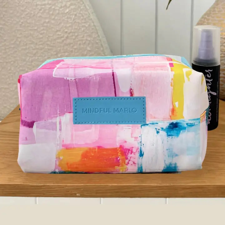 A rectangular cosmetic pouch with a colorful abstract watercolor paint stroke pattern in pink, blue, orange, yellow, and white, featuring a light blue leather patch with the brand name &