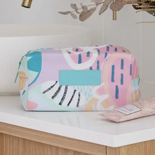 A pastel abstract-print cosmetic pouch sits on a white countertop in a bathroom setting, with a tube of lotion nearby and a sink and dried leaves in the background.