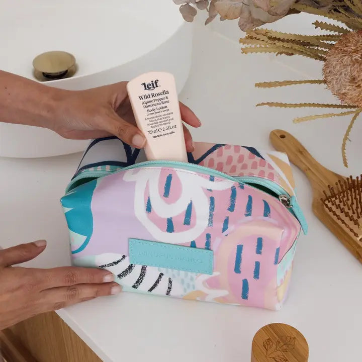 A multicolor cosmetic pouch with a pastel abstract print is shown on a white vanity table, with hands holding a Telf Wild Beauty serum inside it. The background includes a wooden hairbrush and dried flowers.