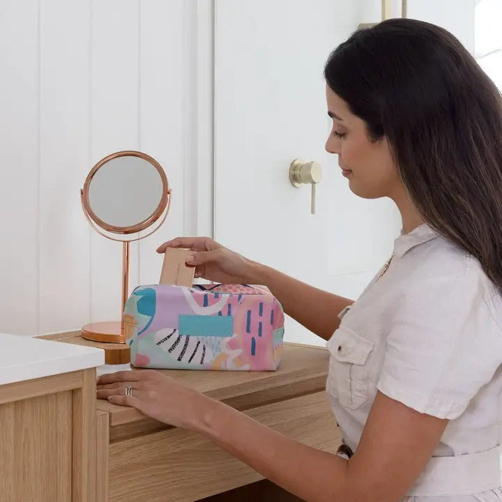 A woman in a white shirt reaches for a colorful tissue box on a wooden vanity table next to a rose gold mirror.