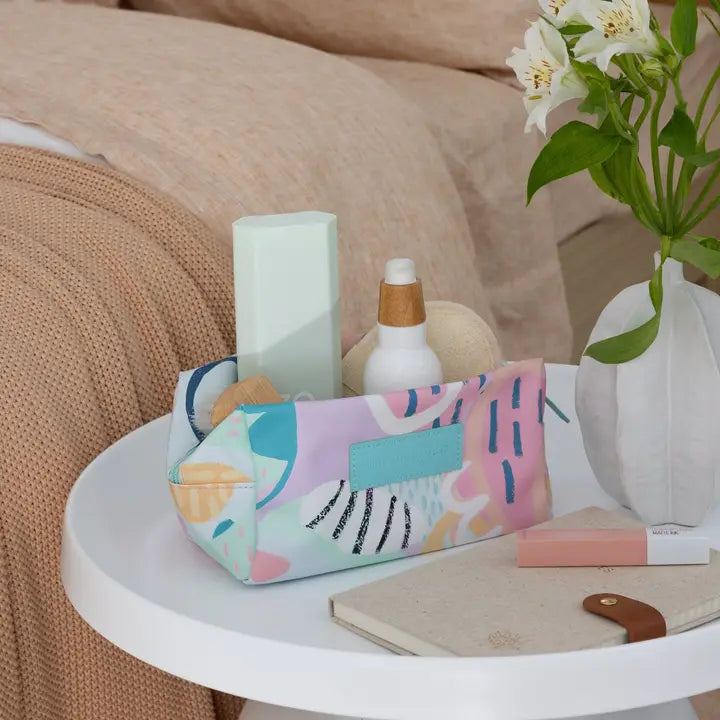 A pastel abstract-pattern makeup bag sits on a white bedside table next to a vase of white flowers, a notebook, and skincare items, in a cozy bedroom setting.