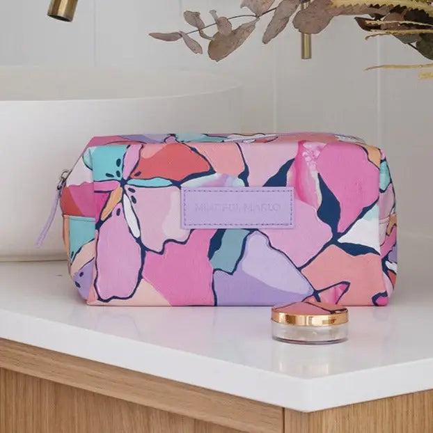 A rectangular makeup bag with a colorful abstract floral pattern in pink, purple, blue, and orange, resting on a white bathroom counter next to a small jar.