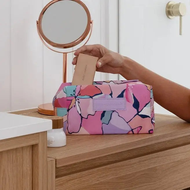 Person using a pink floral makeup bag on a wooden vanity with a mirror.