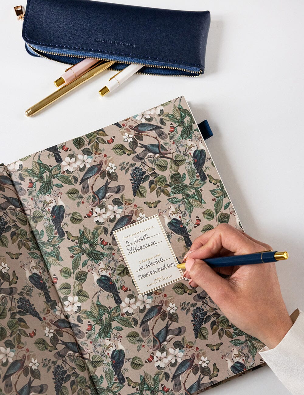 Floral-patterned notebook with a hand holding a pen, navy pencil case, and gold pen on a white background