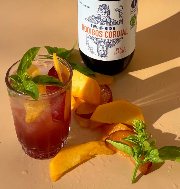 Cocktail with peach slices and basil leaves next to a bottle of Two by Bush Rooibos Cordial on a beige surface.