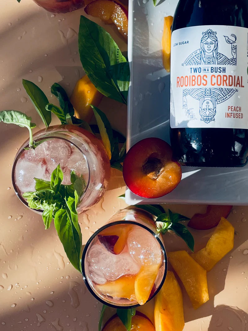 Two Bush Rooibos Cordial bottle with peach slices and mint leaves on a wet surface