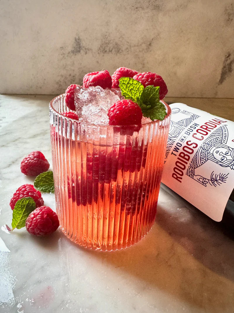 Pink cocktail with raspberries and mint leaves in a ribbed glass, accompanied by a bottle on a marble surface.