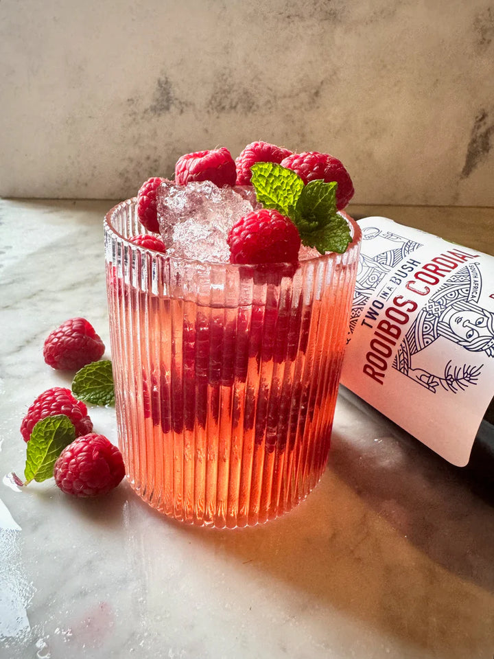 Pink cocktail with raspberries and mint leaves in a ribbed glass, accompanied by a bottle on a marble surface.
