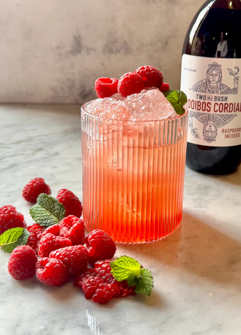 Cocktail with raspberries and a bottle of Two Bush Cordial on a marble surface