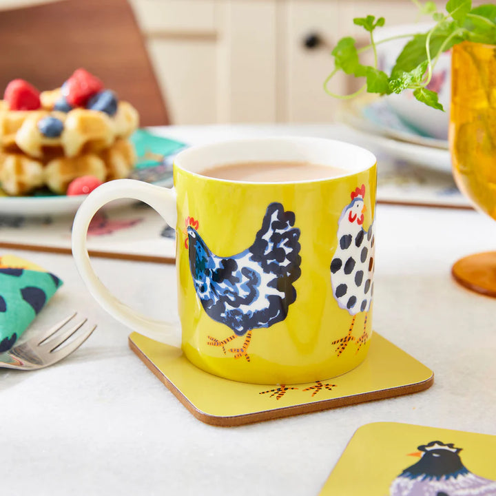 Yellow mug with chicken design on a coaster, placed on a kitchen counter with a plate of waffles and berries in the background.