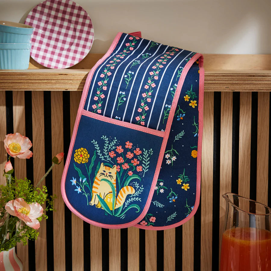 Floral and cat-themed oven mitt hanging on a wooden surface with a checkered plate and flowers in the background.