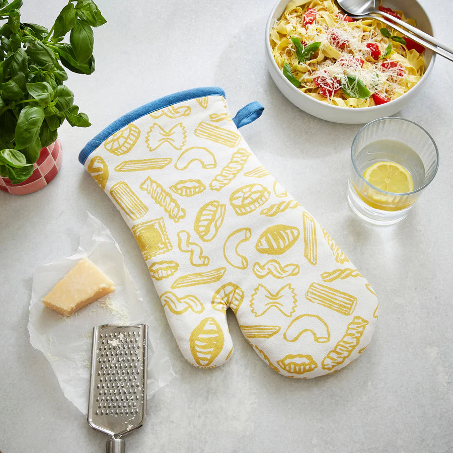 Oven mitt with pasta pattern on a table with pasta dish, glass of water, and grater.
