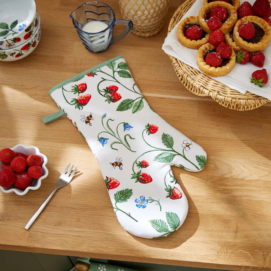 Oven mitt with strawberry pattern on a wooden table with berries and pastries.
