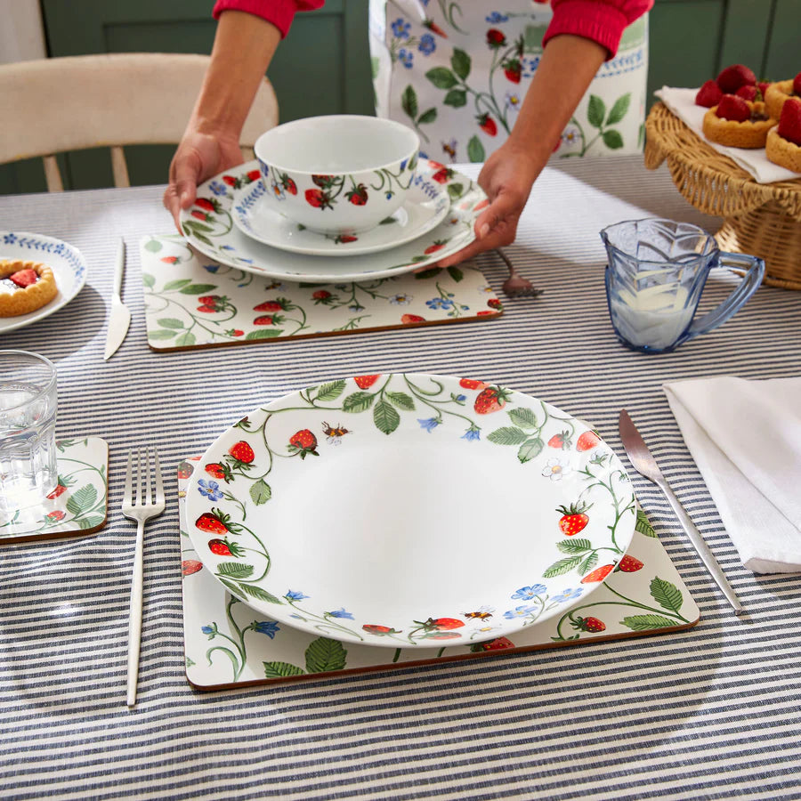 Person setting a table with floral and strawberry-patterned dishes on a striped tablecloth.