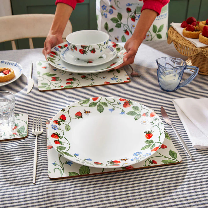 Person setting a table with floral and strawberry-patterned dishes on a striped tablecloth.