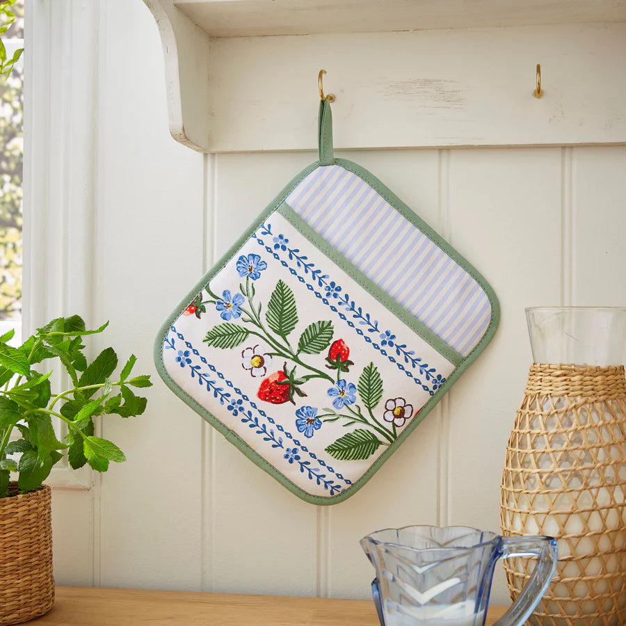 Floral embroidered pot holder hanging on a hook with a plant and vase in the background.