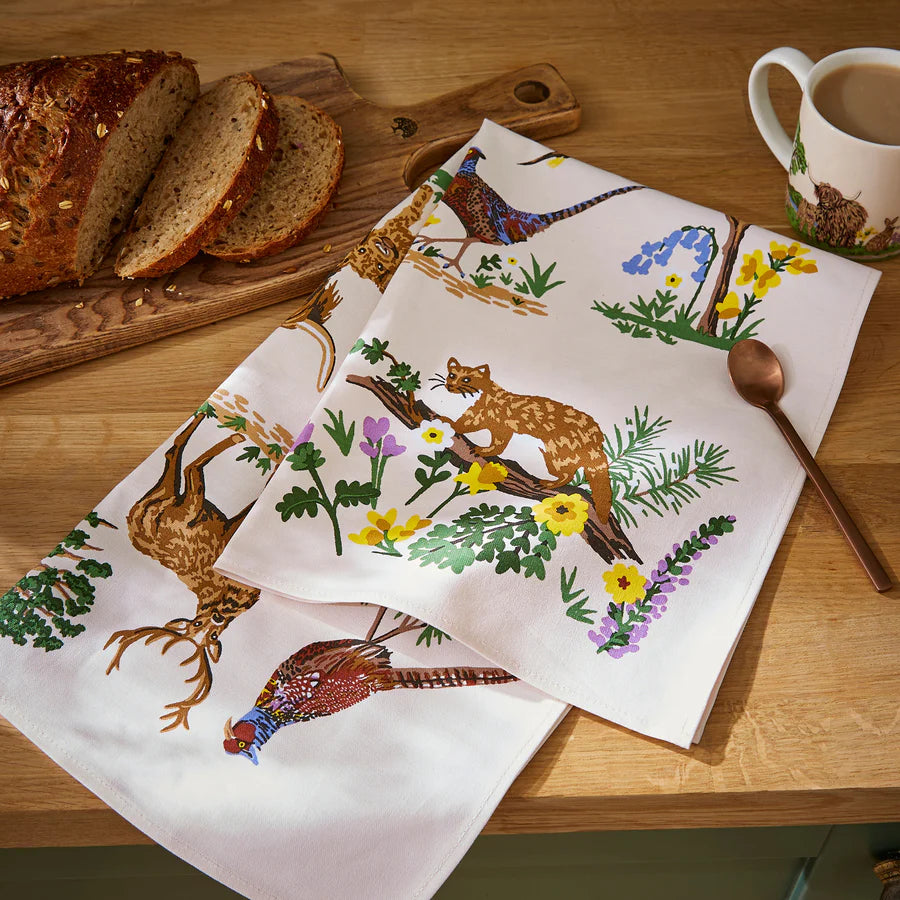 A tea towel with nature-themed design on a wooden table with bread and a mug.
