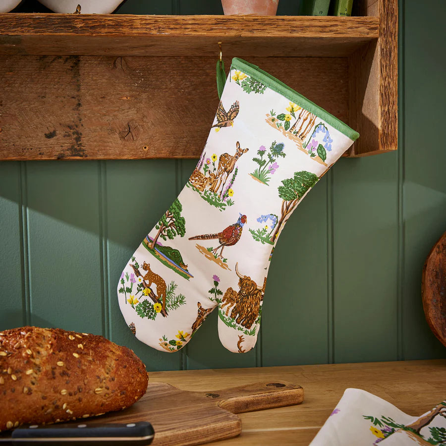 Oven mitt with nature-themed design hanging on a wooden shelf with bread and cutting board below.