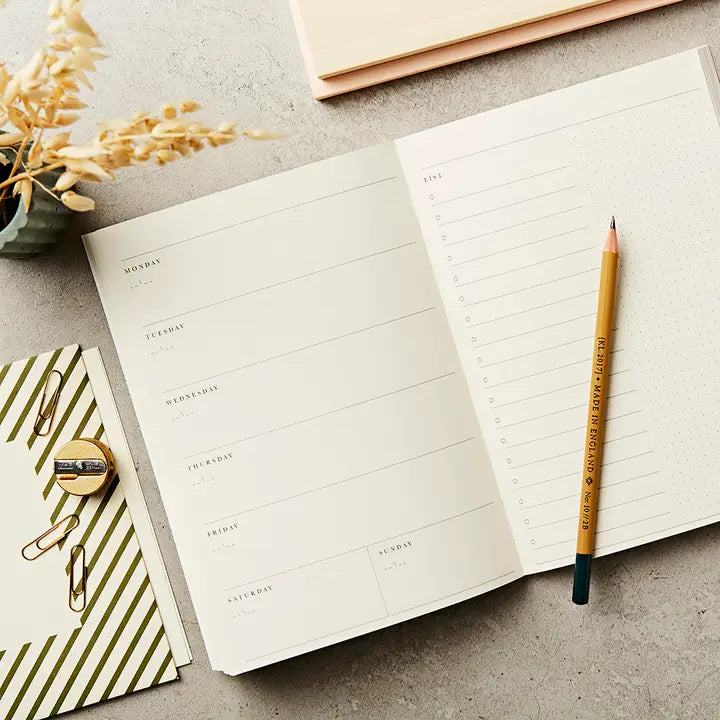 Open planner with a pencil on a concrete surface, surrounded by decorative items.