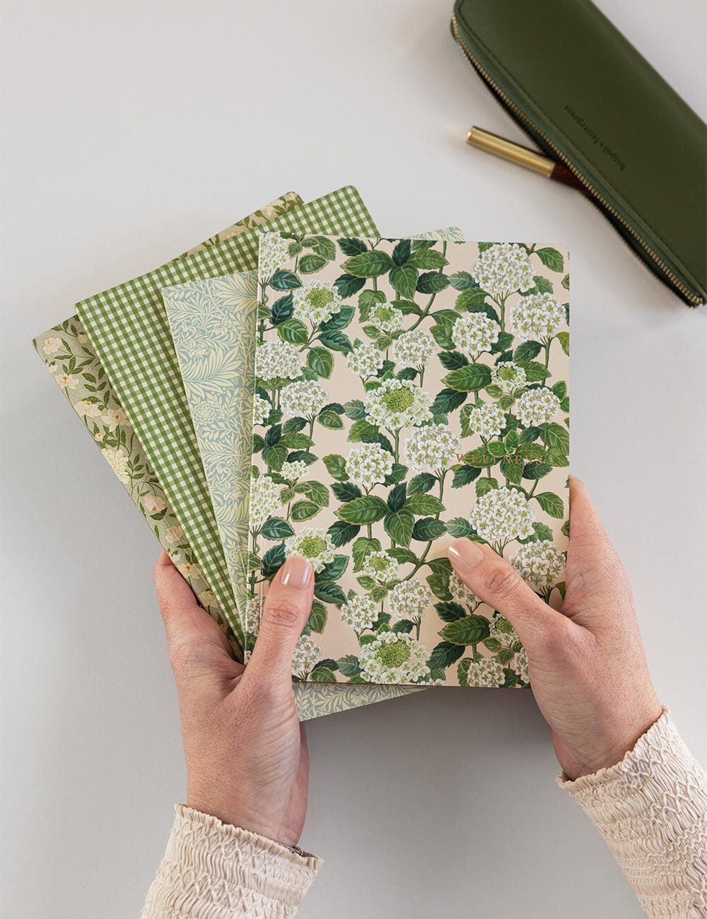 Hands holding three green-themed notebooks with floral and checkered patterns on a light gray background.