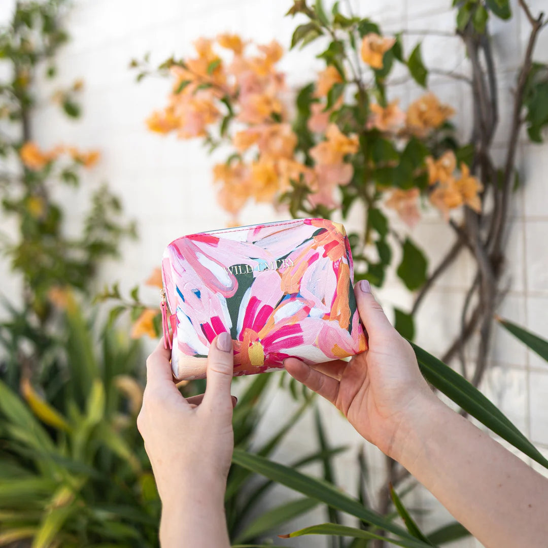 Person holding a colorful floral-patterned cosmetic case in front of plants with flowers.