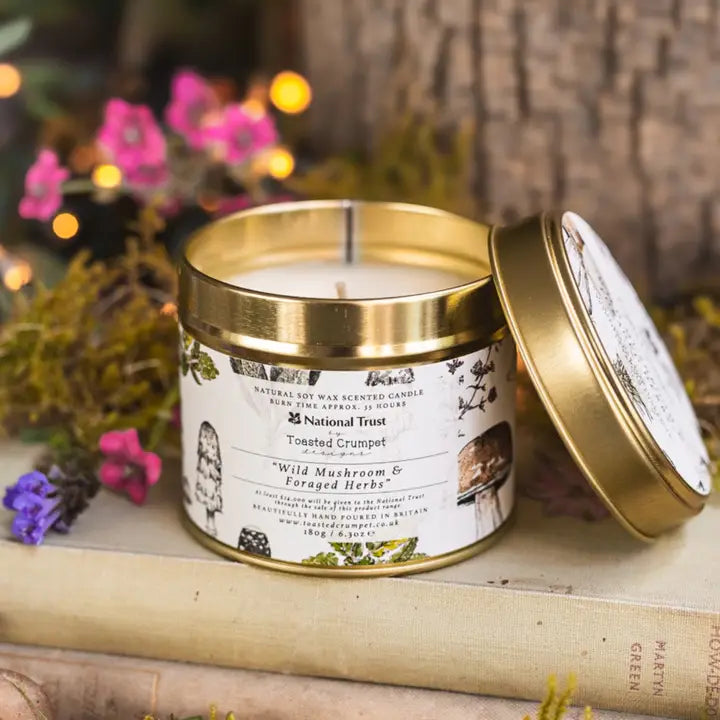 Candle in a metal tin with a National Trust label, surrounded by flowers and books.