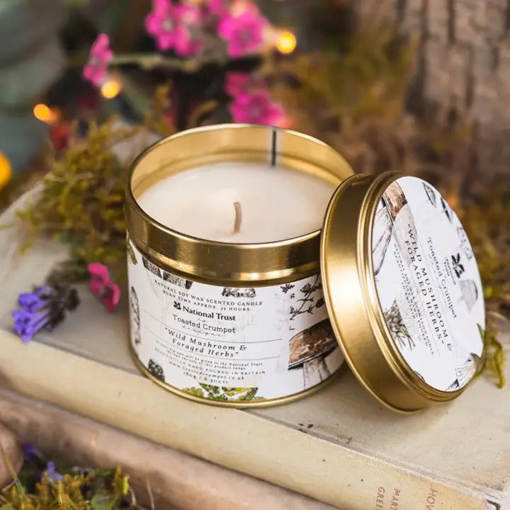 Candle in a gold tin with a National Trust label, surrounded by flowers and natural elements.