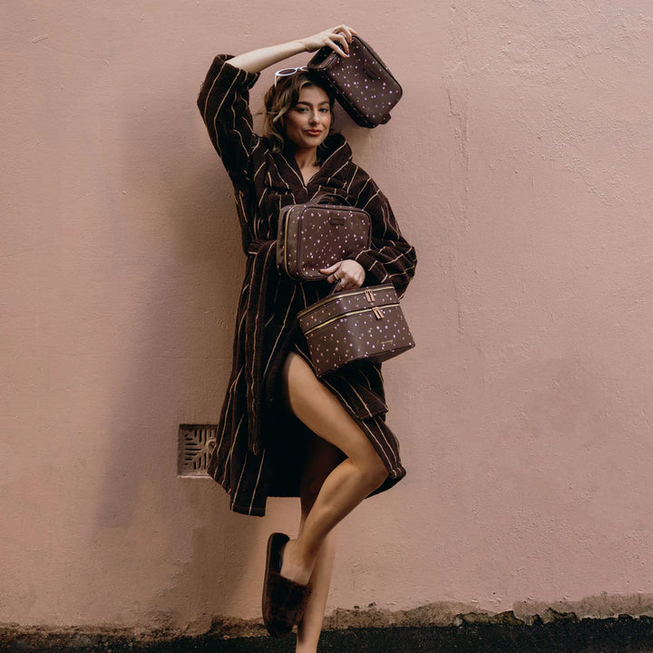 Woman in a dark robe holding patterned bags against a beige wall