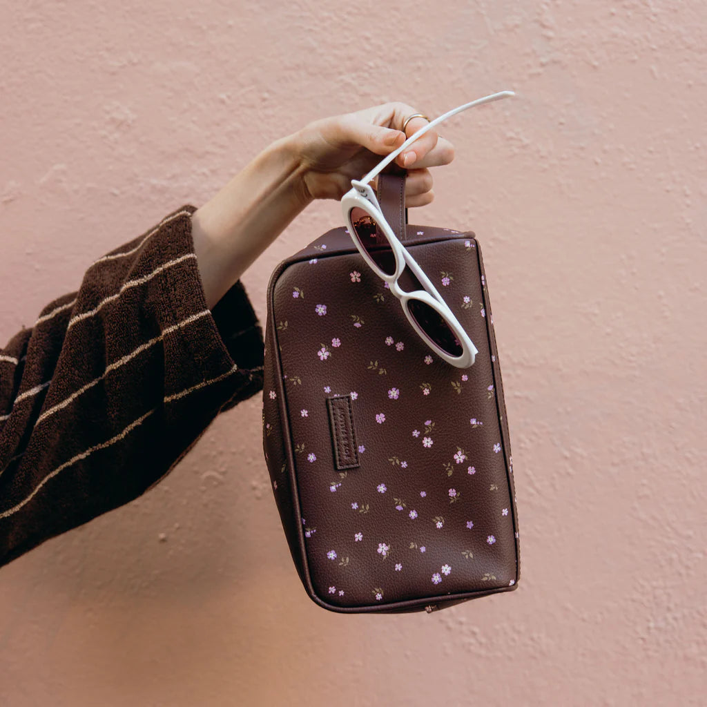 Hand holding a brown floral-patterned pouch with sunglasses on a pink background