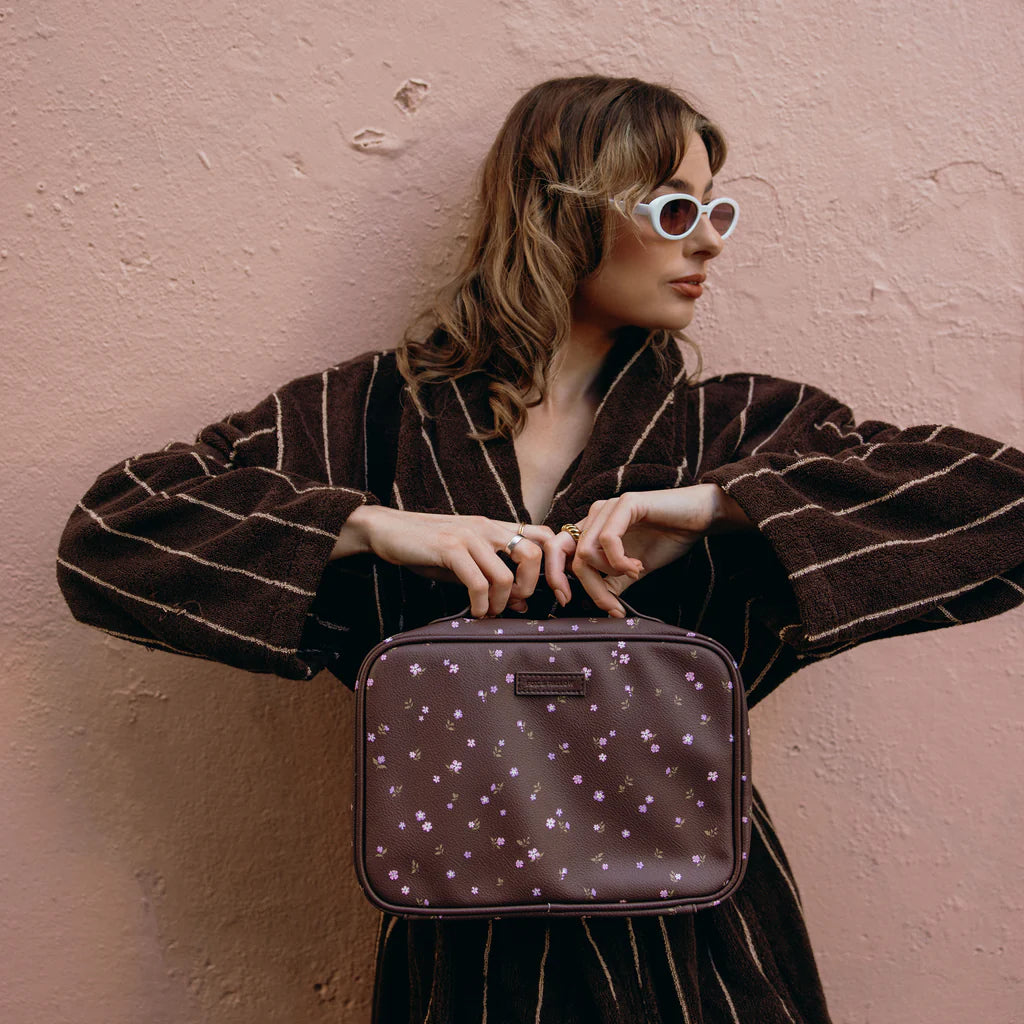 Woman holding a purple floral-patterned cosmetic bag against a pink wall