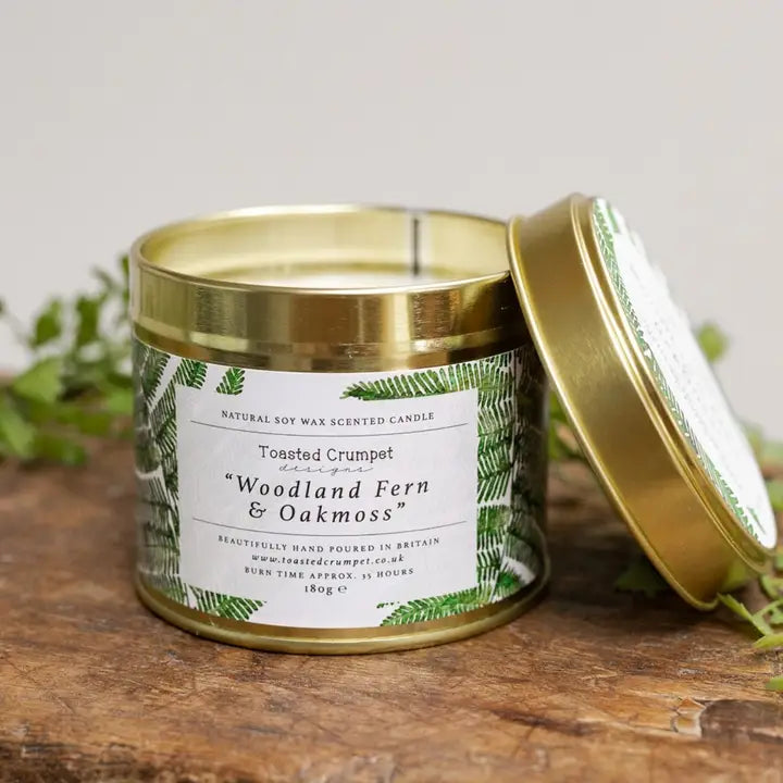 Scented candle with a gold lid and label on a wooden surface, surrounded by greenery.