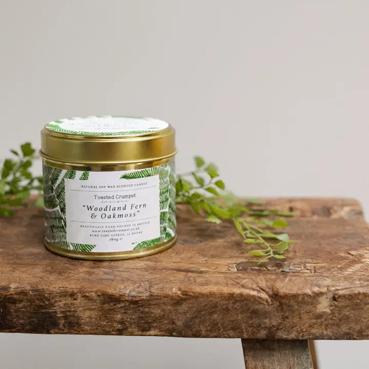 Candle jar labeled 'Woodland Fern & Oakmoss' on a wooden surface with greenery.