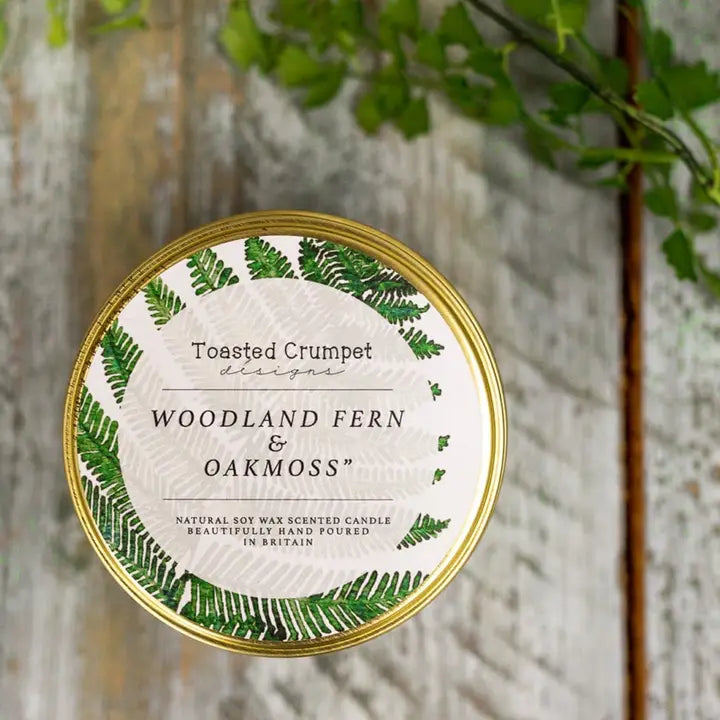 Candle tin labeled 'Woodland Fern & Oakmoss' by Toasted Crumpet Designs on a wooden background.